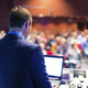 A man is photographed from behind. He is looking on the screen of a laptop. The audience is sitting in the background