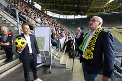 Links steht Christoph Edeler mit BVB-Fußball in der rechten Hand. Rechts steht Rektor Manfred Bayer mit Mikrofon und einem Fanschal, der auf der einen Seite grün mit TU-Logo und rechts schwarz-gelb mit BVB-Logo ist.