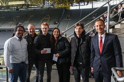 Eine Gruppe von Personen steht vor einer Tribüne gefüllt mit Erstsemesterstudierenden.