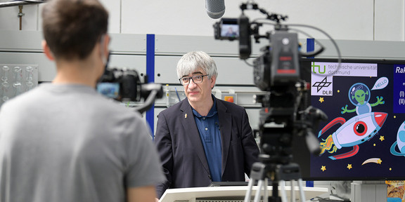 DLR_School_Lab Ein Mann wird von zwei Kameras aufgezeichnet. Er steht hinter einem Pult.