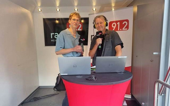 Ein Radiomoderator und ein Schüler stehen mit Mikrofonen in der Hand hinter einem rot betuchten Stehtisch. Im Hintergrund sieht man ein Banner des Radiosenders.