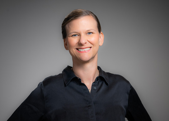 Portrait photo of Prof. Tessa Flatten: Prof. Flatten wears a black blouse and stands in front of a gray background. She smiles at the camera.