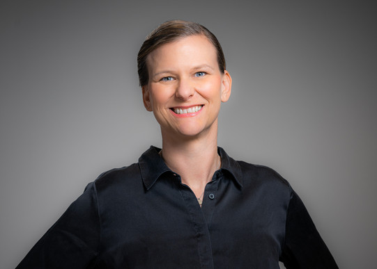 Portrait photo of Prof. Tessa Flatten: Prof. Flatten wears a black blouse and stands in front of a gray background. She smiles at the camera.