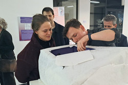 Das Bild zeigt einen weißen Gegenstand mit einem rechteckigen Einschnitt. Zwei Personen schauen in diesen Ausschnitt hinein. Weitere Personen stehen dahinter.