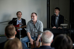 The picture shows a band of three male figures. The backs of several people's heads can be seen in the foreground.