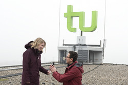 Ein Mann kniet mit einem Bein vor einer Frau und hält eine Ringbox in den Händen. Sie stehen auf dem Dach des Mathetowers vor dem TU-Logo.