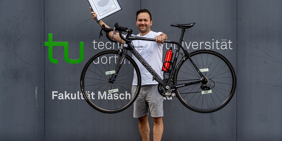 Ein Portrait von Dr. Dennis Freiburg, der mit seinem Fahrrad und einer Urkunde vor einem Gebäude der TU Dortmund steht.