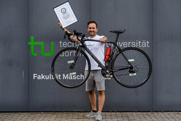 Ein Portrait von Dr. Dennis Freiburg, der mit seinem Fahrrad und einer Urkunde vor einem Gebäude der TU Dortmund steht.