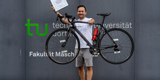 Ein Portrait von Dr. Dennis Freiburg, der mit seinem Fahrrad und einer Urkunde vor einem Gebäude der TU Dortmund steht.