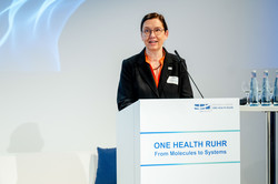 Foto: Prof. Barbara Albert,Rektorin der Universität Duisburg-Essen, steht an einem Redepult. Albert trägt eine Brille, einen schwarzen Blazer und eine rote Bluse.