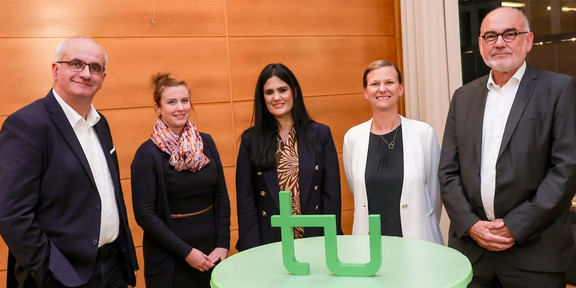 Zwei Männer und drei Frauen stehen um einen Stehtisch. Auf dem Stehtisch steht ein grüner TU-Logo-Aufsteller.