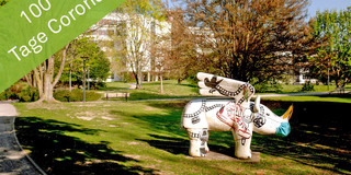 Eine große, weiße Skulptur eines geflügelten Nashorns, dem ein Mundschutz angelegt wurde, steht auf einer Wiese. Im Hintergrund sind große, weiße Gebäude zu sehen.