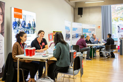 Gespräche auf der Messe In einem großen Raum stehen viele Tische, an denen Menschen Gespräche führen