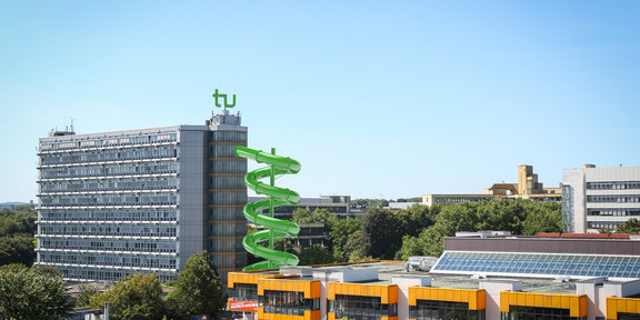 Rutsche Mathetower Bearbeitetes Bild: Blick auf die Mensabrücke mit Mathetower und Mensagebäude. An dem Mathetower, ausgehend vom 9. Stockwerk, ist eine grüne, gewundene Rutsche zu sehen.