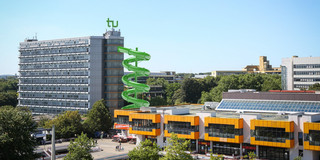 Rutsche Mathetower Bearbeitetes Bild: Blick auf die Mensabrücke mit Mathetower und Mensagebäude. An dem Mathetower, ausgehend vom 9. Stockwerk, ist eine grüne, gewundene Rutsche zu sehen.