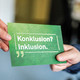 Eine Postkarte mit der Aufschrift "Konklusion? Inklusion!" wird von einer Hand zur anderen gereicht.