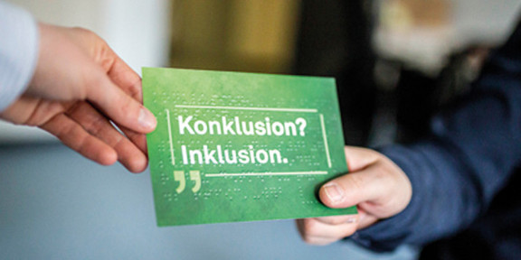 Eine Postkarte mit der Aufschrift "Konklusion? Inklusion!" wird von einer Hand zur anderen gereicht.