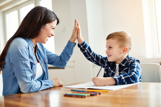 Mutter und Kind geben sich ein High-Five am Küchentisch, während das Kind Hausaufgaben macht