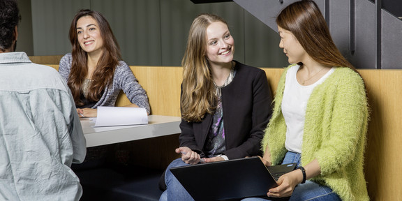Drei Frauen und ein Mann sitzen an einem Tisch und unterhalten sich. Eine der Frauen hält einen Laptop in der Hand.