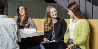 Drei Frauen und ein Mann sitzen an einem Tisch und unterhalten sich. Eine der Frauen hält einen Laptop in der Hand.