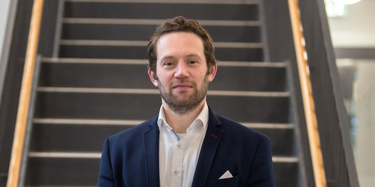 Porträtfoto von Prof. Sascha Alavi: Prof. Alavi trägt einen dunklen Anzug und steht mit gefalteten Händen vor einer Treppe.