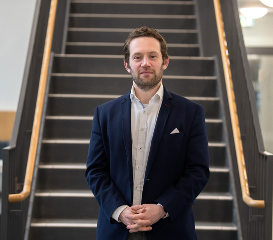 Porträtfoto von Prof. Sascha Alavi: Prof. Alavi trägt einen dunklen Anzug und steht mit gefalteten Händen vor einer Treppe.