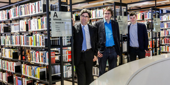 Die Verantwortlichen der Bibliotheken-Allianz stehen vor Bücherregalen der Universitätsbibliothek.