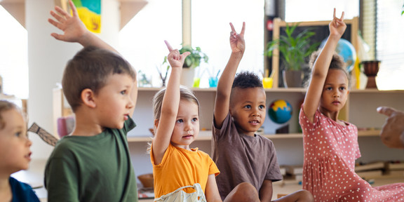 Foto: Vorschulkinder sitzen auf dem Boden eines Kita-Raums und heben die Finger hoch.