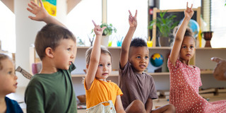Foto: Vorschulkinder sitzen auf dem Boden eines Kita-Raums und heben die Finger hoch.