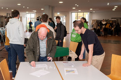 Um Tische im Audimax-Foyer stehen Besucher*innen und Studierende. Vorne ist ein älterer Herr zu sehen, der würfelt. Neben ihm steht ein Student, der etwas erklärt.