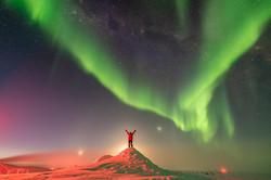 A man in red jacket on a small snow hill under a night sky with green auroras and numerous stars.