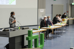 Multiple people with face masks sit at separate tables in front of a screen. A woman is standing behind a lectern.