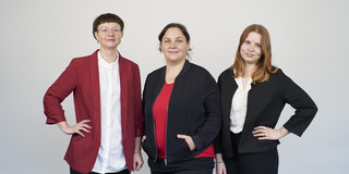 Das Bild zeigt drei nebeneinanderstehende Frauen vor einem weißen Hintergrund
