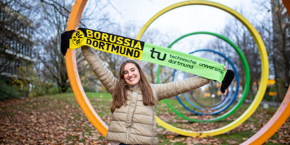 A young woman holds up a scarf with the BVB and TU Dortmund University logos in front of the spectral rings on the TU campus