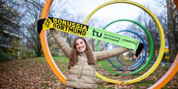 Eine junge Frau hält einen Schal mit den Logos des BVB und der TU Dortmund vor den Spektralringen auf dem TU-Campus in die Luft