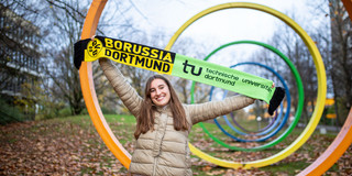 Eine junge Frau hält einen Schal mit den Logos des BVB und der TU Dortmund vor den Spektralringen auf dem TU-Campus in die Luft
