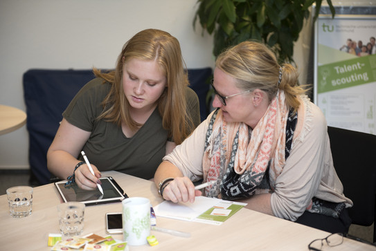 Zwei Frauen sitzen an einem Tisch und sprechen miteinander. Eine Frau schreibt auf einem Tablet und die andere hat einen Stift in der Hand.