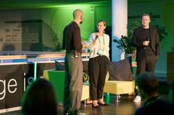 Foto: Drei Personen stehen auf der Bühne, zwei von ihnen halten Mikrofone und sprechen miteinander.