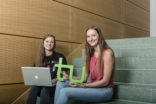 Zwei Studentinnen der TU Dortmund sitzen auf einer Treppenstufe mit einem Laptop und dem TU Logo. 