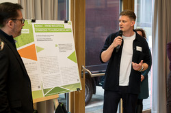 A young blond man is presenting a poster and speaking into a microphone. Other people are standing around him.