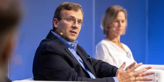 A man with glasses and a suit sits in a white armchair and speaks into a portable microphone. In the background, a woman is also sitting in a white armchair. 