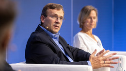 Ein Mann mit Brille und Anzug sitz in einem weißen Sessel und spricht in ein tragbares Mikrofon. Im Hintergrund sitzt eine Frau ebenfalls in einem weißen Sessel. 
