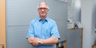 Portrait von Prof. Christian Bühler. Seine Haare sind kurz und grau. Er trägt eine schwarze Brille. Er steht mit verschränkten Armen im hellblauen Hemd vor einer Bürowand und lächelt in die Kamera.