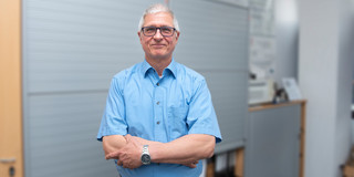 Portrait of Prof. Christian Bühler. His hair is short and gray. He wears black glasses. He is standing in front of an office wall with his arms folded in a light blue shirt and smiling at the camera.