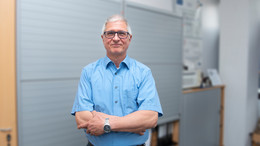 Portrait von Prof. Christian Bühler. Seine Haare sind kurz und grau. Er trägt eine schwarze Brille. Er steht mit verschränkten Armen im hellblauen Hemd vor einer Bürowand und lächelt in die Kamera.