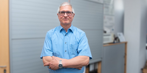 Prof. Christian Bühler Portrait von Prof. Christian Bühler. Seine Haare sind kurz und grau. Er trägt eine schwarze Brille. Er steht mit verschränkten Armen im hellblauen Hemd vor einer Bürowand und lächelt in die Kamera.