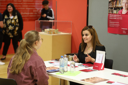Zwei junge Frauen sitzen sich an einem Tisch gegenüber und führen ein Beratungsgespräch. Auf dem Tisch liegen Informationsmaterialien und im Hintergrund sind weitere Menschen sowie Plakate zu sehen.