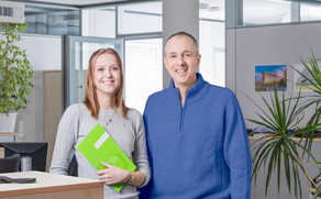 A woman with a green folder in her hand and a man are standing in an office and smiling at the camera.
