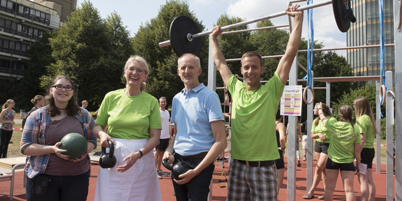 TUrnpunkt Zwei Frauen und zwei Männer halten Sportgeräte in den Händen, der rechte Mann stemmt eine Langhantel, im Hintergrund ist der TUrnpunkt zu sehen