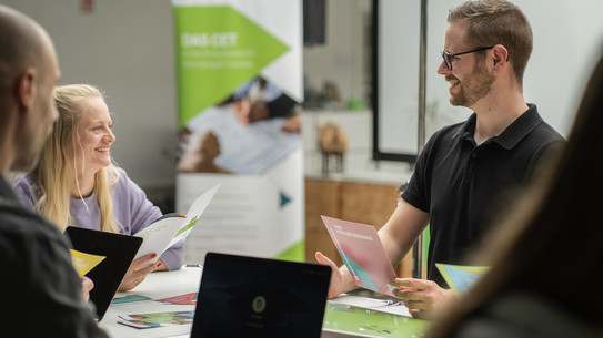 Mehrere Personen sitzen an einem Tisch mit Laptop und Broschüren und besprechen Informationsmaterial in einem hellen Raum.